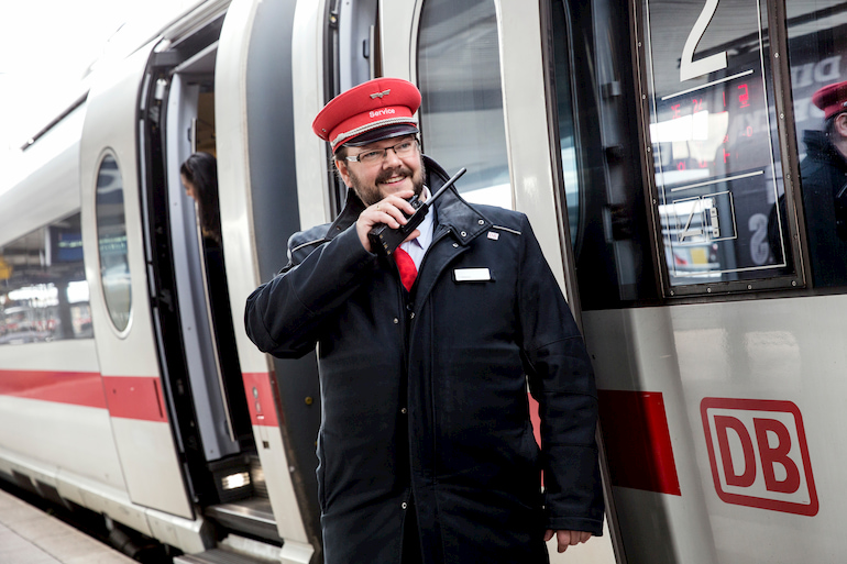 Ein Mann in Deutscher-Bahn-Uniform spricht vor einem Zug in ein Walkie-Talkie.