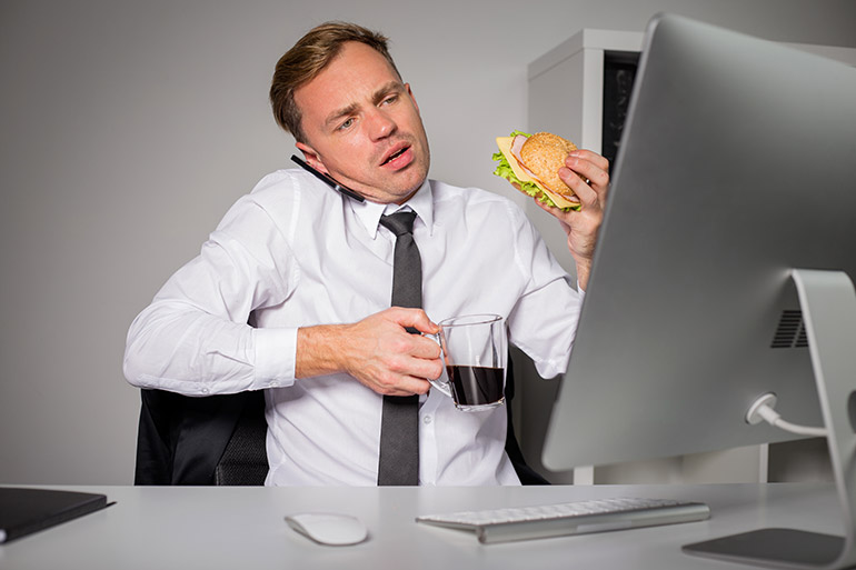 Ein Mann isst Brötchen und trinkt Kaffee, während er am Bildschirm sitzt.