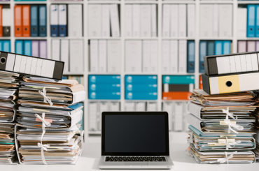 Ein Notebook auf einem Schreibtisch, der mit vielen Akten und Dokumenten zugestapelt ist.
