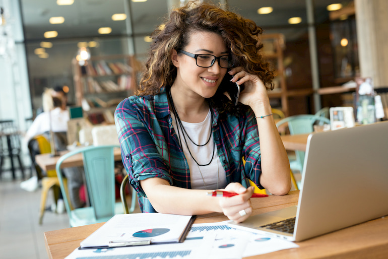 Eine junge Frau sitzt in einem Café vor ihrem Laptop und Arbeitspapieren, während die telefoniert.