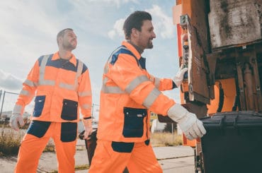 Zwei Männer tragen Arbeitskleidung und arbeiten bei der Müllabfuhr.