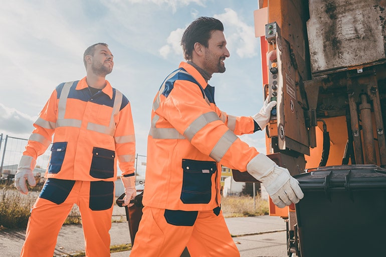 Zwei Männer tragen Arbeitskleidung und arbeiten bei der Müllabfuhr.