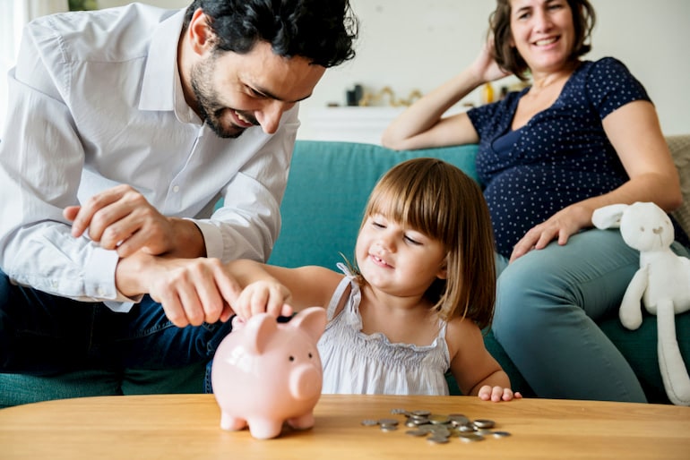 Mutter und Vater sehen Kind dabei zu, wie es Geld in ein Sparschwein tut.