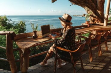 Eine junge Frau arbeitet mit Blick aufs Meer im Homeoffice im Ausland.