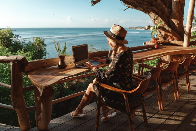 Eine junge Frau arbeitet mit Blick aufs Meer im Homeoffice im Ausland.